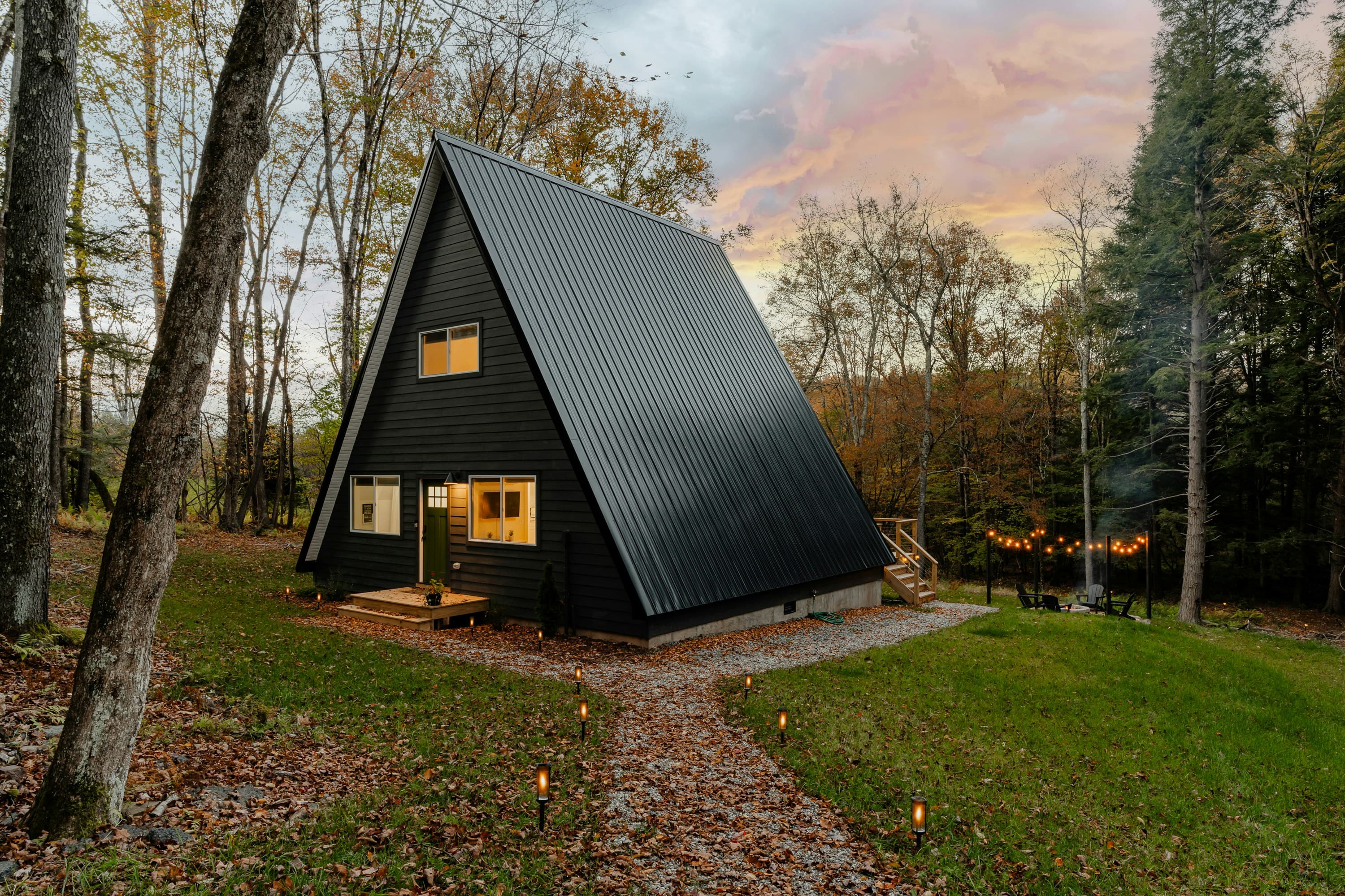 Dark A-frame cabin among autumn trees, with a gravel path leading to the entrance at dusk